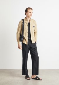 Camicia button-up beige con dettaglio della tasca, T-shirt nera, pantaloni neri a gamba larga e sandali neri. Il modello sta in piedi con una borsa a spalla.