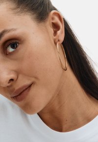 Gold hoop earrings feature a smooth, circular design that reflects light. The model wears a white shirt, highlighting the earrings' shine.