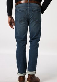 Vaqueros de mezclilla en azul oscuro, con corte recto, dobladillos remangados y dos bolsillos traseros. Llevados con botas de cuero marrón y una camisa negra.