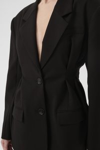 Svart blazer med dyp V-hals, strukturerte skuldre, to frontlommer og en enkelt knappelukkning, laget av et glatt stoff.