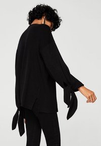 Pull noir à manches longues avec des accents de nœud aux poignets et des fentes sur les côtés. Texture douce et coupe décontractée, mettant en valeur un design simple et moderne.