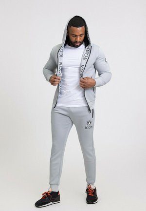 Ensemble de sport gris clair comprenant un sweat à capuche zippé et un pantalon assorti. Présente des accents de logo et un t-shirt blanc, porté avec des baskets noires.