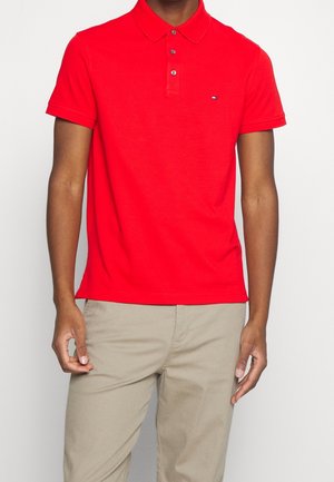 Czerwona koszulka polo wykonana z bawełny, z klasycznym kołnierzykiem, trzema guzikami oraz małym logo na piersi. Noszona z jasnobeżowymi spodniami.