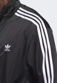 Czarna kurtka na zamek z gładkiego materiału. Posiada biały logo Adidas oraz trzy białe paski biegnące wzdłuż każdego ramienia. Wysoki kołnierz dodatkowo podkreśla detal.