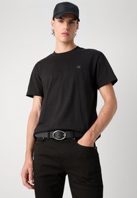 T-shirt in cotone nero con collo rotondo e maniche corte. Presenta un piccolo logo CK ricamato sul petto. Indossata con jeans neri e una cintura.