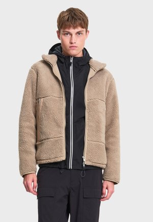 Jeune homme portant une veste polaire beige sur un sweat à capuche noir zippé et un pantalon noir, sur un fond clair uni.