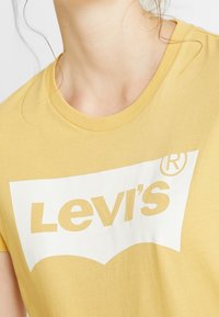 T-shirt z krótkim rękawem w kolorze żółtym, wykonany z bawełny, z białym logo z napisem "Levi's". Gładka tekstura i okrągły dekolt, casualowy design.