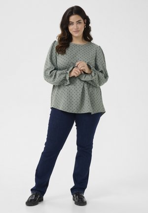 Blouse verte à pois avec des manches bouffantes, associée à un jean bleu foncé et des chaussures noires. La blouse présente un col rond et des poignets froncés.