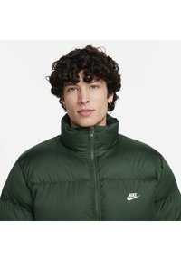 Ciemnozielona kurtka puchowa z wysokim kołnierzem, zapięciem na zamek oraz białym logo Nike po lewej stronie klatki piersiowej. Gładka, wyściółkowana tekstura z wypukłymi szwami.