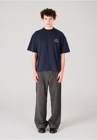 Camiseta de algodón azul marino con mangas cortas y cuello redondo; presenta un pequeño logo dorado. Combinada con pantalones grises de corte holgado y zapatos negros.