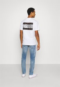 Homem de costas, vestindo uma camisola branca gráfica da Tommy Jeans, jeans azuis claros e ténis brancos de cano alto, encostado a um fundo simples.