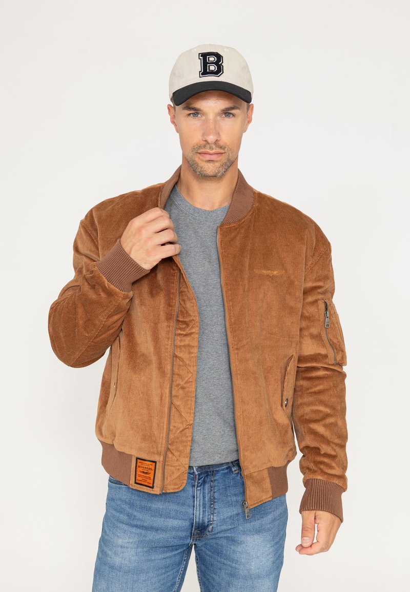 Bruine corduroy bomberjack met ribgebreide manchetten, zijzakken en een oranje label. Draagt over een grijs t-shirt en gecombineerd met blauwe spijkerbroeken.