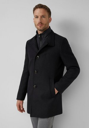 Man die een zwarte wollen jas met knopen draagt over een zwarte jas met rits, staand met één hand in zijn zak tegen een grijze achtergrond.