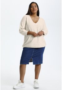 Beige gebreide trui met een V-hals en lange mouwen, gecombineerd met een donkerblauwe denim kokerrok met een voorknof. Witte sneakers.