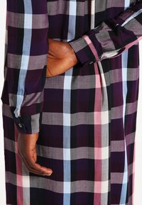 Personne portant une chemise à manches longues à carreaux violets, bleus, roses, noirs et blancs, une main glissée derrière le dos.