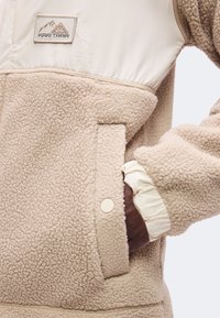 Veste en polaire sherpa beige avec poche à boutons-pression et poignet élastique, avec un écusson logo montagne sur la poitrine.