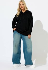 Felpa nera oversize con cappuccio e cordoncino, abbinata a jeans in denim azzurro chiaro a gamba larga con effetto sbiadito. Sneakers bianche.