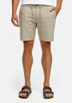 Homme portant un short beige mi-genou avec cordon de serrage et des sandales noires à boucles, debout avec une main dans la poche devant un fond uni.