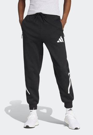Osoba nosząca czarne spodnie dresowe Adidas z białym logo i paskami, białe skarpetki i białe buty sportowe, stojąca z rękami w kieszeniach.