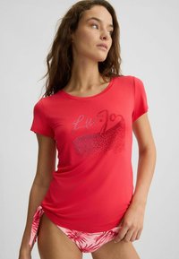 Raudona trumpa rankovė į T-shirt su apvaliu iškirpimu, papuošta blizgančiu logotipo dizainu. Suderinta su rožinėmis gėlėmis raštuotomis maudymosi kelnaitėmis.