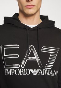 Joven con sudadera negra con gran logo blanco de EA7 Emporio Armani y franjas estilizadas en la parte frontal.