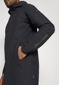 Giacca impermeabile nera con cappuccio, tasche laterali con zip, strisce riflettenti sulle maniche e un dettaglio verde all'orlo.