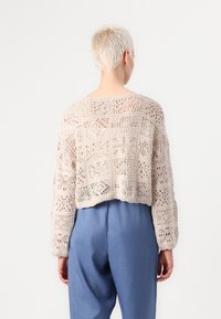 Ljust beige virkat tröja med öppet mönster, kort längd och långa ärmar, kombinerad med blå högmidjade byxor.