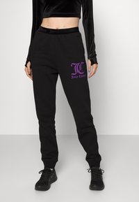 Černé tepláky s elastickým pasem, obsahující boční kapsy a fialové logo "Juicy Couture" na horní levé nohavici.