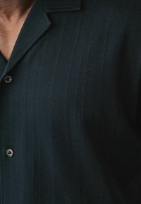 Camicia nera con un motivo a spina di pesce testurizzato, dotata di colletto a revers e due bottoni visibili vicino al collo.