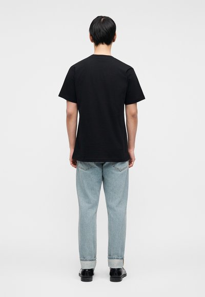 Μαύρο βαμβακερό μπλουζάκι με κοντά μανίκια, από πίσω. Φορεμένο με ανοιχτό μπλε τζιν και μαύρα παπούτσια, με διπλωμένα μανίκια.