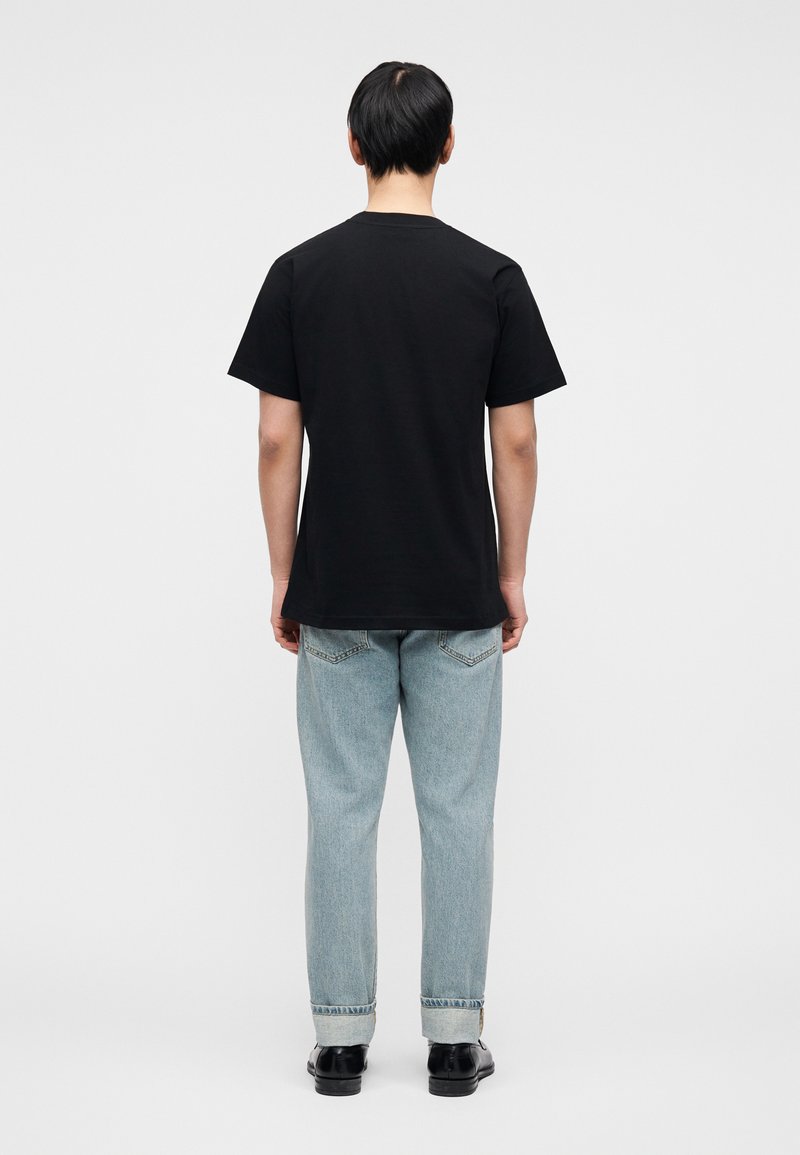 T-shirt en coton noir à manches courtes, vu de dos. Porté avec un jean en denim bleu clair et des chaussures noires, avec des revers de pantalon.