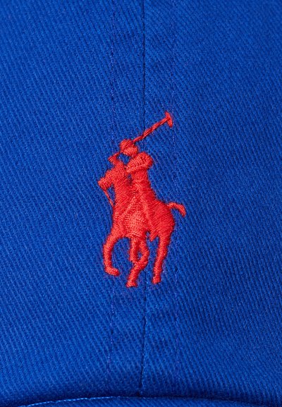 Mėlynas kepuraitė su ryškiu raudonu siuvinėtu polo žaidėjo logotipu. Audinys turi smulkų tekstūrinį raštą.