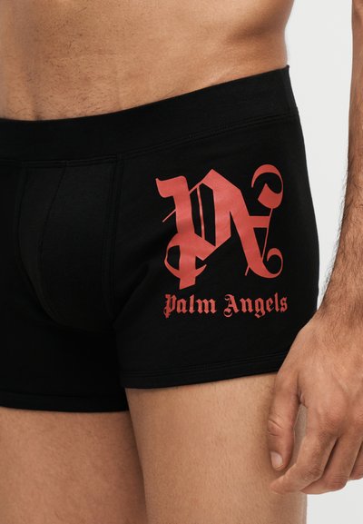 Άνδρας φοράει μαύρα boxer briefs με κόκκινο λογότυπο "Palm Angels" στο αριστερό μηρό και το χέρι ακουμπάει δίπλα.