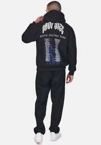 Schwarzer Hoodie mit großem weißen Text auf dem Rücken, der die Tourorte auflistet. Das Material wirkt weich; kombiniert mit schwarzen Hosen und weißen Turnschuhen.