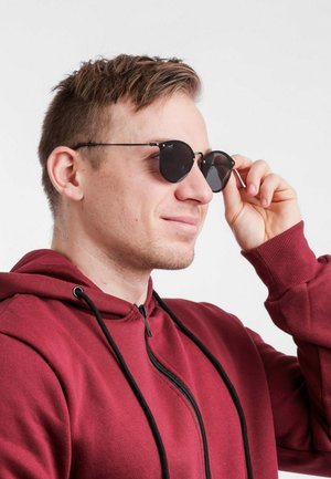 Kulaté černé sluneční brýle s tmavými skly, kovový rám a tenké nožičky. Nosí je osoba v vínové mikině na zip, která si brýle nastavuje.