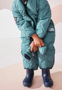 Tâchê gewatteerde jumpsuit in teal met een rits, voorzien van een текстур patroon en mouwen met duimgaten; gecombineerd met marineblauwe rubberen laarzen.