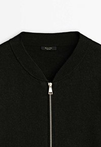 Veste noire à col côtelé avec une fermeture éclair argentée et une petite étiquette de marque à l'intérieur du col.