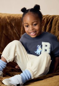 Grau Sweatshirt mit einem "B" und einem Cartoon-Hundedesign, cremefarbene Jogginghosen, blaue gerippte Socken und weiße Sneaker. Weiche Texturen und lässige Passform.