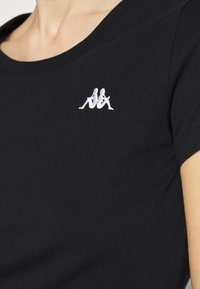 Černé bavlněné tričko s krátkým rukávem, se stříbrně vyšitým logem na hrudi. Tkanina má hladkou strukturu.