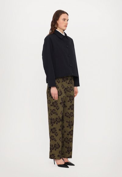 Marineblauw button-up jasje met een kraag, gecombineerd met olijfgroene broek met bloemenpatroon en wijdere benen, en zwarte puntige hakken.