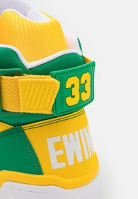Gros plan sur le talon d'une chaussure de sport verte et jaune avec "33" sur la sangle et "EWING" en lettres blanches sur fond jaune.