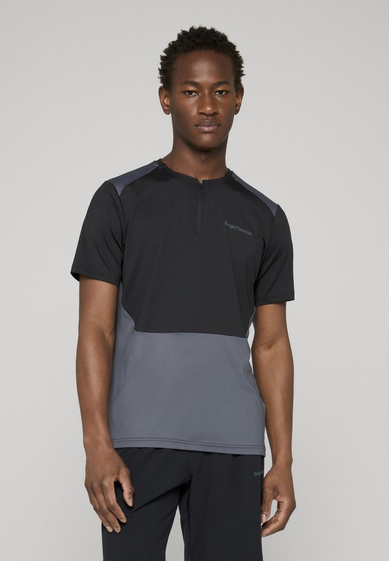 Jeune homme portant un t-shirt de sport à manches courtes noir et gris avec une fermeture éclair à l'avant, debout devant un fond uni gris clair.