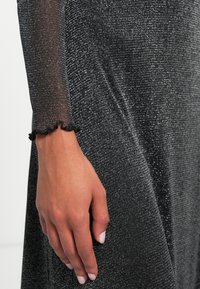 Fekete, csillogó ruha texturált anyagból. Hosszú, áttetsző ujja, fekete, fodros mandzsettával. Közeli kép egy kézről, amely a ruhán nyugszik.