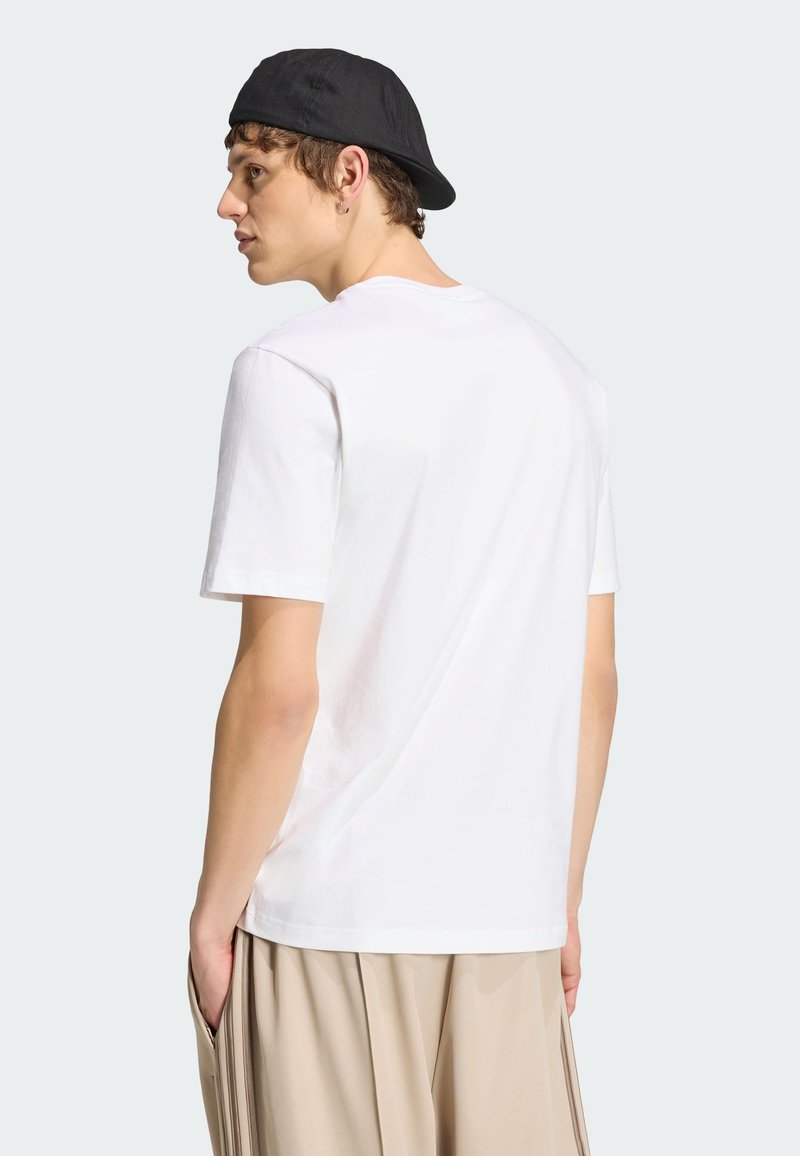 Junger Mann in weißem T-Shirt, beigen Hosen und schwarzer rückwärts getragener Kappe, steht mit Händen in den Taschen, nach links blickend, vor hellem Hintergrund.