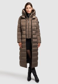 Ilgas rudas pūkuotas paltas su kapišonu, su siuvinėta danga, priekinis užtrauktukas ir šoniniai kišeniai, derinamas su juoda megztine suknele ir ilgais juodais botais.