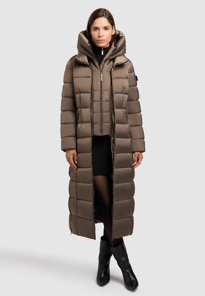 Ilgas rudas pūkuotas paltas su kapišonu, su siuvinėta danga, priekinis užtrauktukas ir šoniniai kišeniai, derinamas su juoda megztine suknele ir ilgais juodais botais.