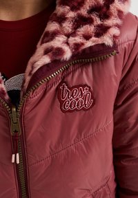 Röd vadderad jacka med en rosa fluffig krage, med ett broderat märke som säger "tres cool" och detaljer i brons på dragkedjan. Slät textur och kviltad design.