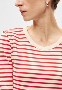 Πολυάριθμο μπλουζάκι με λεπτές κόκκινες και κρεμ οριζόντιες ρίγες, με στρογγυλή λαιμόκοψη και ελαφρώς εφαρμοστή γραμμή για μια μοντέρνα εμφάνιση.