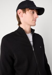 Sweat à fermeture éclair noir avec un col haut, présentant une texture lisse et un logo brodé sur la poitrine. Associé à une casquette noire.