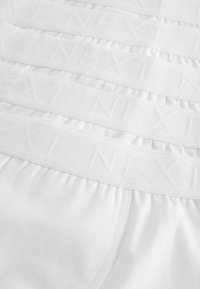 Culottes en coton blanc avec une taille élastique douce, arborant des lettres blanches « NEXT » en relief. Cinq paires empilées horizontalement.